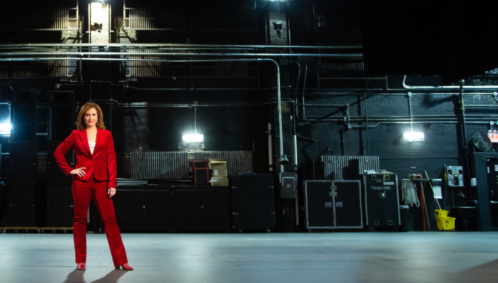 Tricia Brouk stands on stage in a red suit and with stage lights in the backdrop